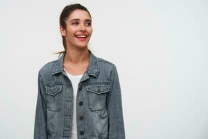 Portrait of young smiling brunette lady wears in white t-shirt and denim jackets, looks away with happy expression, stands over white background with copy space on the right side. photo