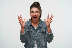 Photo of young aggressive brunette lady wears in white t-shirt and denim jackets, screaming and looks angry with raised hands, stands over white background.