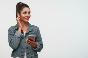 Young cheerful brunette lady wears in white t-shirt and denim jackets, holds smartphone and broadly smiles, listening cool song ant toches new headphones. photo