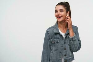 Young positive brunette lady wears in white t-shirt and denim jackets, looks away and broadly smiles, listening cool song in headphones, stands over white background wit copy space. photo