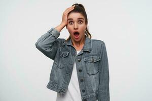 Photo of young shocked brunette lady wears in white t-shirt and denim jackets, looks at the camera with hwide open mouth and eyes, keep palm on the head, stands over white background.