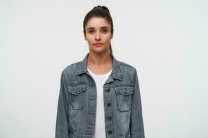 Portrait of young brunette lady wears in white t-shirt and denim jackets, looks at the camera with calm expression, stands over white background. photo
