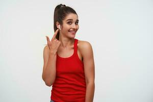 Portrait of young cheerful brunette lady wears in red t-shirt, looks at the camera and broadly smiles, listening cool song and pointing at headphones, stands over white background with copy space. photo