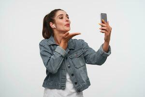 Young cheerful brunette lady wears in white t-shirt and denim jackets, holds smartphone and sends kiss to the videochat. Stands over white background. photo