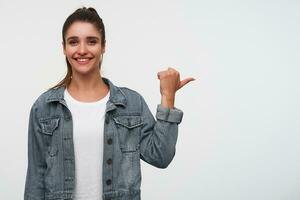 Young cheerful brunette lady wears in white t-shirt and denim jackets, looks at the camera and broadly smiles, points fingers to the right side on copy space, stands over white background. photo