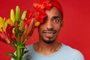 joven loco atractivo hombre en rojo sombrero y azul camiseta, sostiene un ramo de flores en su manos y cubierto parte de cara con flores, mira a el cámara con amplio abierto ojos, soportes terminado rojo fondo foto