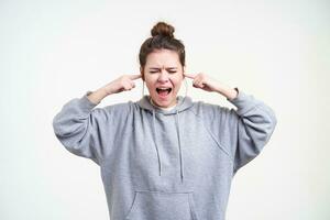 Stressed young brunette lady with bun hairstyle shouting with closed eyes and inserting forefingers into ears, standing over white background in sporty wear photo