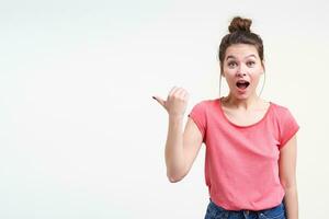 Excited young pretty brunette woman with bun hairstyle raising surprisedly eyebrows while showing emotionally aside with thumb, isolated over white background photo