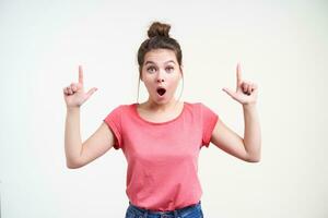 Excited young pretty long haired female with bun hairstyle keeping her hands raised while showing dazedly upwards with forefingers, standing over white background photo