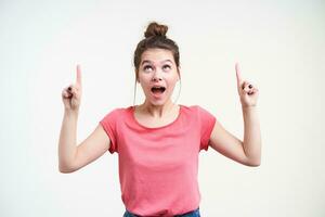 Agitated young lovely brown haired female keeping her mouth opened while pointing excitedly upwards with index fingers, isolated over white background photo