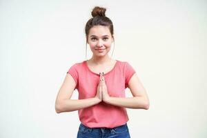 Charming young attractive blue-eyed brunette lady with bun hairstyle raising hands in praying gesture and smiling pleasantly to camera while posing over white background photo