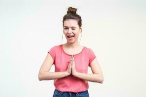 Joyful young lovely brunette female keeping one eye closed while looking playfully at camera and folding raised hand in praying gesture, isolated over white background photo