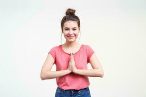 Cheerful young pretty brown haired lady keeping raised palms together while looking gladly at camera with wide smile, standing over white background in casual wear photo