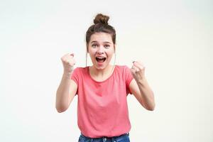 Joyful young pretty brunette female with bun hairstyle raising happily fists while looking excitedly at camera with opened mouth, standing over white background photo