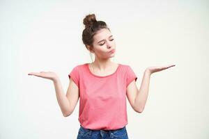 Puzzled young pretty woman wearing her brown hair in knot while posing over white background, keeping palms raised and looking pensively on one of it photo