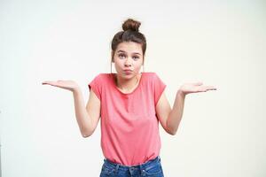 Bewildered young blue-eyed brunette female with natural makeup shrugging with raised palms and looking confusedly at camera, isolated over white background photo