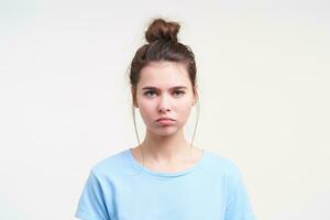 Upset young lovely brunette woman wearing her brown hair in knot while posing over white background, keeping her lips folded while looking sadly at camera photo