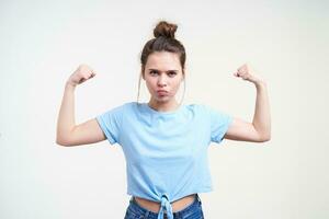 Severe young blue-eyed brown haired woman with bun hairstyle looking at camera with serious face while demonstrating her power with raised hands, standing over white background photo