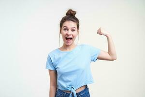 Excited young attractive dark haired woman with bun hairstyle keeping her hand raised while demonstrating her strong biceps, isolated over white background photo