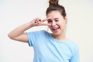 Cheerful young pretty brown haired woman with bun hairstyle keeping peace sign near her face and giving gladly wink at camera, isolated over white background photo