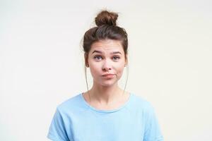 Portrait of young puzzled brown haired lady with bun hairstyle twisting her mouth while looking perplexedly at camera, standing over white background photo