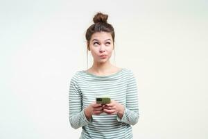 Puzzled young attractive brown haired lady pouting her lips while looking pensively upwards, keeping mobile phone in raised hands while posing over white background photo