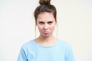 Close-up of offended young blue-eyed brunette lady pouting her lips while looking sadly at camera, dressed in blue t-shirt while standing over white background photo