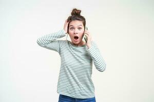 Amazed young pretty brown haired woman with casual hairstyle keeping raised palm on her head while looking dazedly at camera, standing over white background with mobile phone in raised hand photo