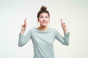 Indoor shot of young attractive brunette female with bun hairstyle raising hands with crossed fingers and looking worringly upwards, isolated over white background photo