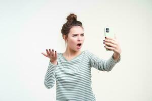 Studio photo of young brown haired indignant lady raising perplexed her palm and frowning eyebrows while having emotional phone talk, standing over white background