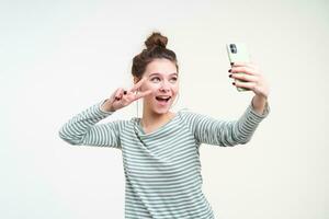 Cheerful young blue-eyed lovely brunette female with bun hairstyle showing peace sign with raised hand while taking selfie with her smartphone, isolated over white background photo