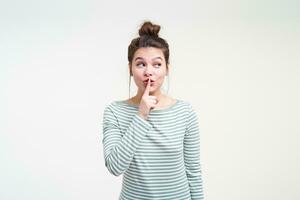 Studio photo of young lovely brunette female wearing her brown hair in knot while posing over white background, holding forefinger on her lips while asking to keep her secret