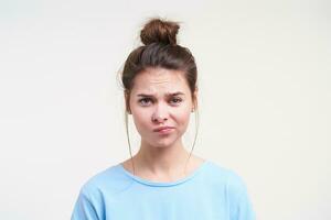 retrato de joven desconcertado bonito marrón peludo dama ceñudo su Cejas mientras mirando confusamente a cámara, en pie terminado blanco antecedentes en azul ropa foto