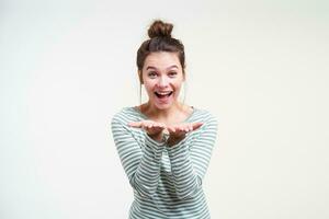 Happy young beautiful brunette woman with bun hairstyle keeping raised palms in front of herself and looking joyfully at camera, standing over white background photo