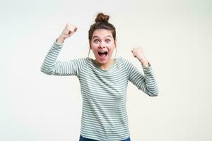 Overjoyed young lovely blue-eyed brown haired female raising emotionally her fists while rejoicing about something, isolated against white background photo