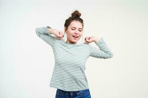 Sleepy young lovely brunette female with natural makeup keeping eyes closed while stretching herself with raised hands, isolated over white background in striped blouse photo