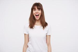 Open-eyed young pretty brown haired lady with casual hairstyle looking excitedly at camera with wide eyes and mouth opened, standing over white backgrounnd photo