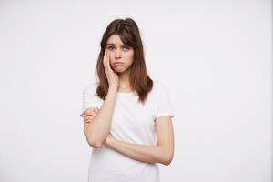 Unhappy young lovely dark haired lady dressed in casual clothes holding hand on her face while looking at camera with upset face, keeping lips folded while posing over white background photo