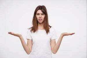 Displeased young pretty dark haired lady with natural makeup raising confusedly her palms while looking at camera with puzzled face, isolated over white background photo