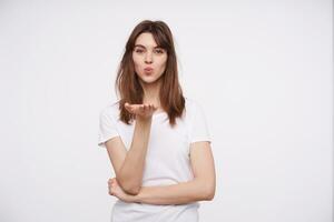 retrato de hermosa joven positivo oscuro peludo dama con casual peinado plegable su labios y enviando aire Beso a cámara, acuerdo palma elevado mientras posando terminado blanco antecedentes foto