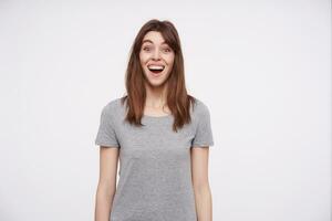 Agitated young lovely brown haired female with natural makeup looking joyfully at camera with wide mouth and eyes opened, isolated over white background photo