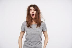 Indoor photo of young pretty tired brown haired woman with wild hair closing her eyes while yawning with wide mouth opened, isolated over white background