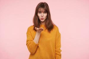 Charming young blue-eyed brunette female folding her lips in air kiss and blowing it at camera, keeping her palm raised while standing over pink background photo