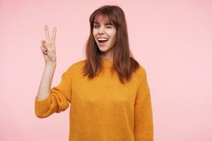 Joyful young pretty dark haired lady with casual hairstyle raising hand with victory gesture and giving wink at camera while smiling widely, standing over pink background photo