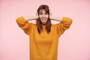 Positive young blue-eyed dark haired lady covering her ears with raised palms and looking cheerfully at camera, sticking out her tongue while posing over pink background photo