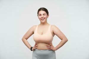 Cheerful young dark haired plump lady in sports bra and leggins keeping hands on waist and winking happily at camera while standing over white background photo