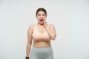 Indoor shot of amazed young oversized female dressed in sporty clothes raising emotionally hand to her face and looking dazedly at camera, isolated over white background photo