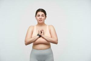 Studio shot of young lovely dark haired oversized woman with casual hairstyle keeping folded hands on her chest and looking hopefully at camera, posing over white background photo