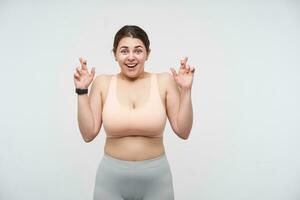 Positive young pretty brunette chubby lady with ponytail hairstyle keeping her fingers crossed while looking cheerfully at camera, standing over white background. Body positive concept photo