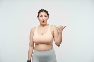 Agitated young dark haired brunette plump lady in sporty clothes looking dazedly at camera with opened mouth and showing aside with thumb, isolated over white background photo